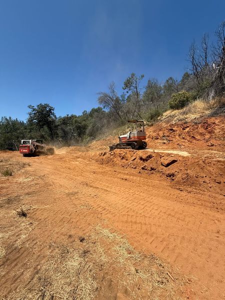 Two orange machines clearing a dirt hillside, kicking up dust under a blue sky.