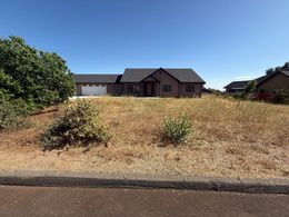 A one-story home with a dark roof, set on a hill with dry grass, under a blue sky.