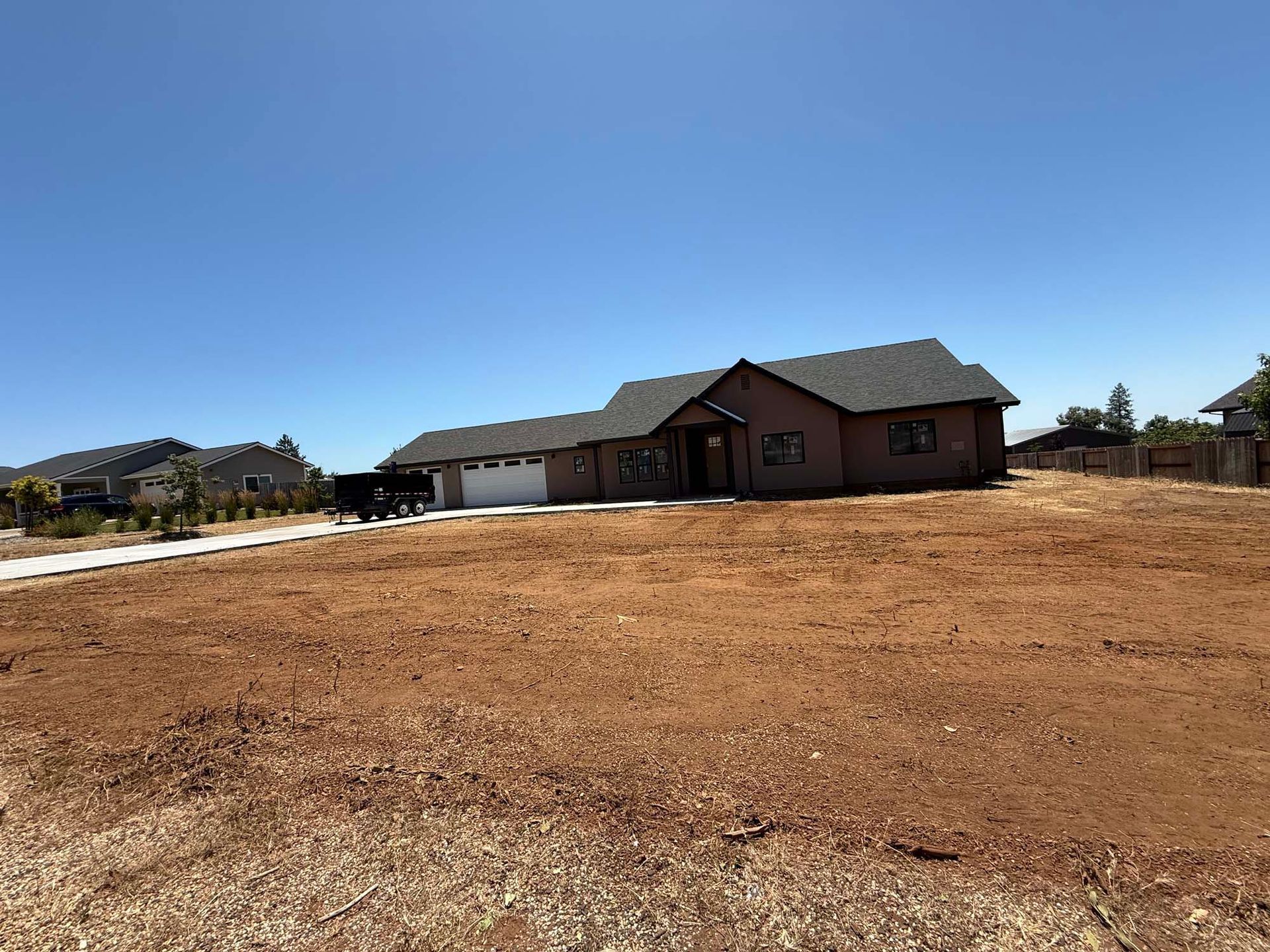 Single-story house on a dirt lot under a blue sky. Brown siding, dark roof, white garage door.
