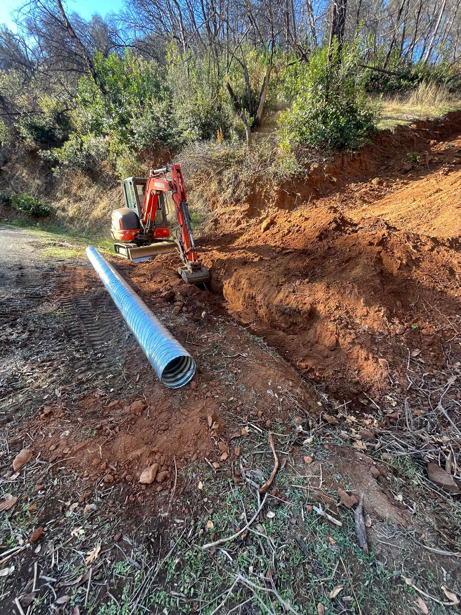 An excavator digs a trench next to a metal pipe, in a dirt hillside. Forest in the background.