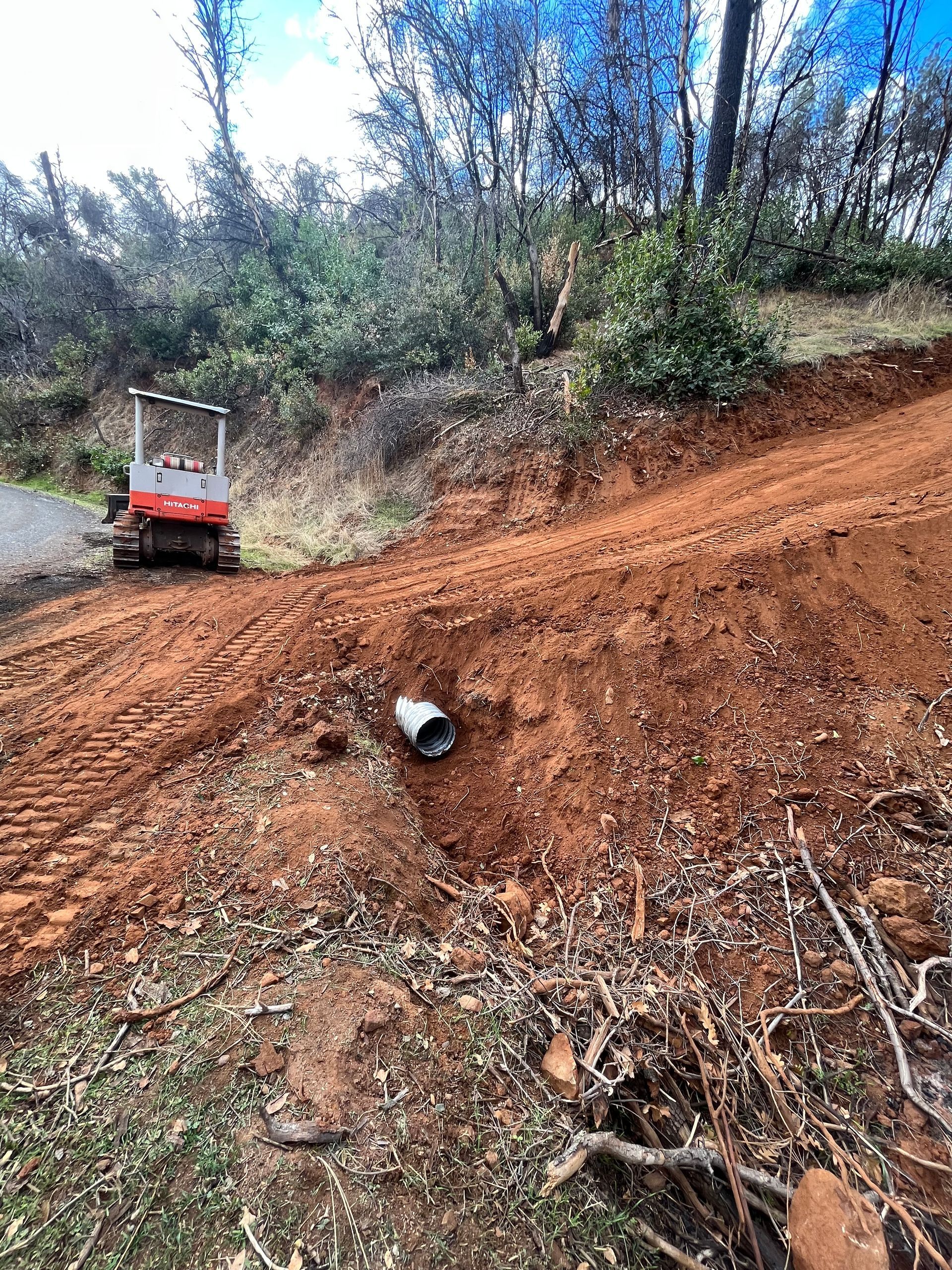 A small excavator on a hillside, red soil, and a drainage pipe visible.