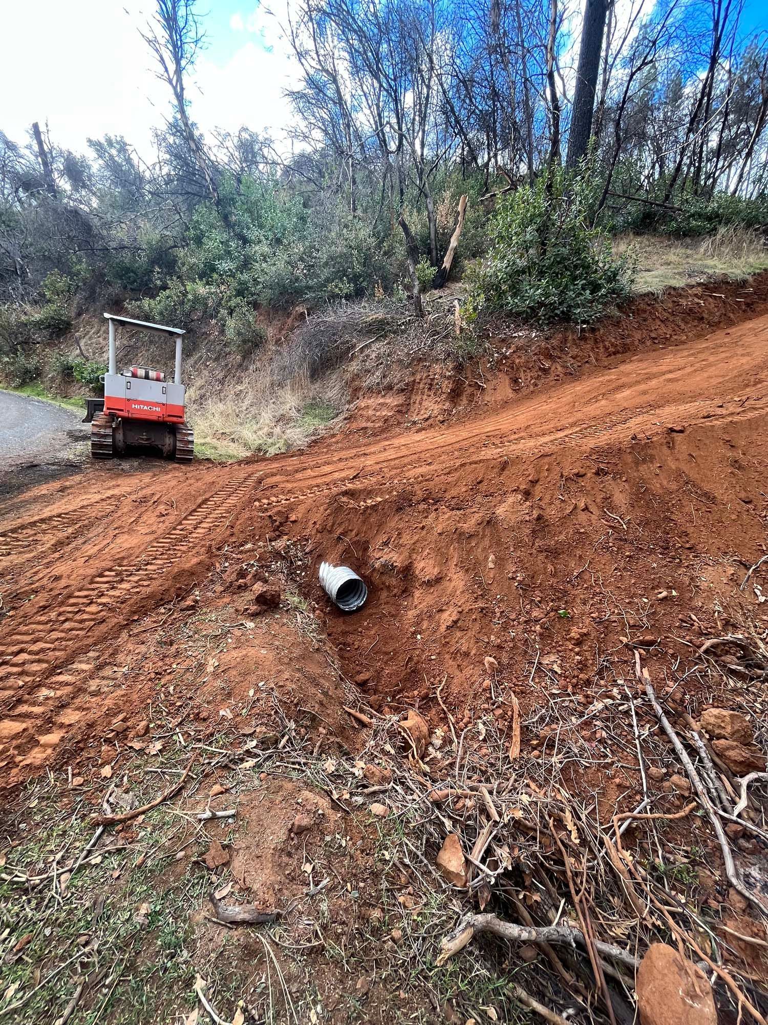 A small excavator works on a dirt hillside with a culvert. Red dirt and forest visible.