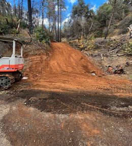 Dirt road construction with small excavator on a hillside, trees in background under blue sky.