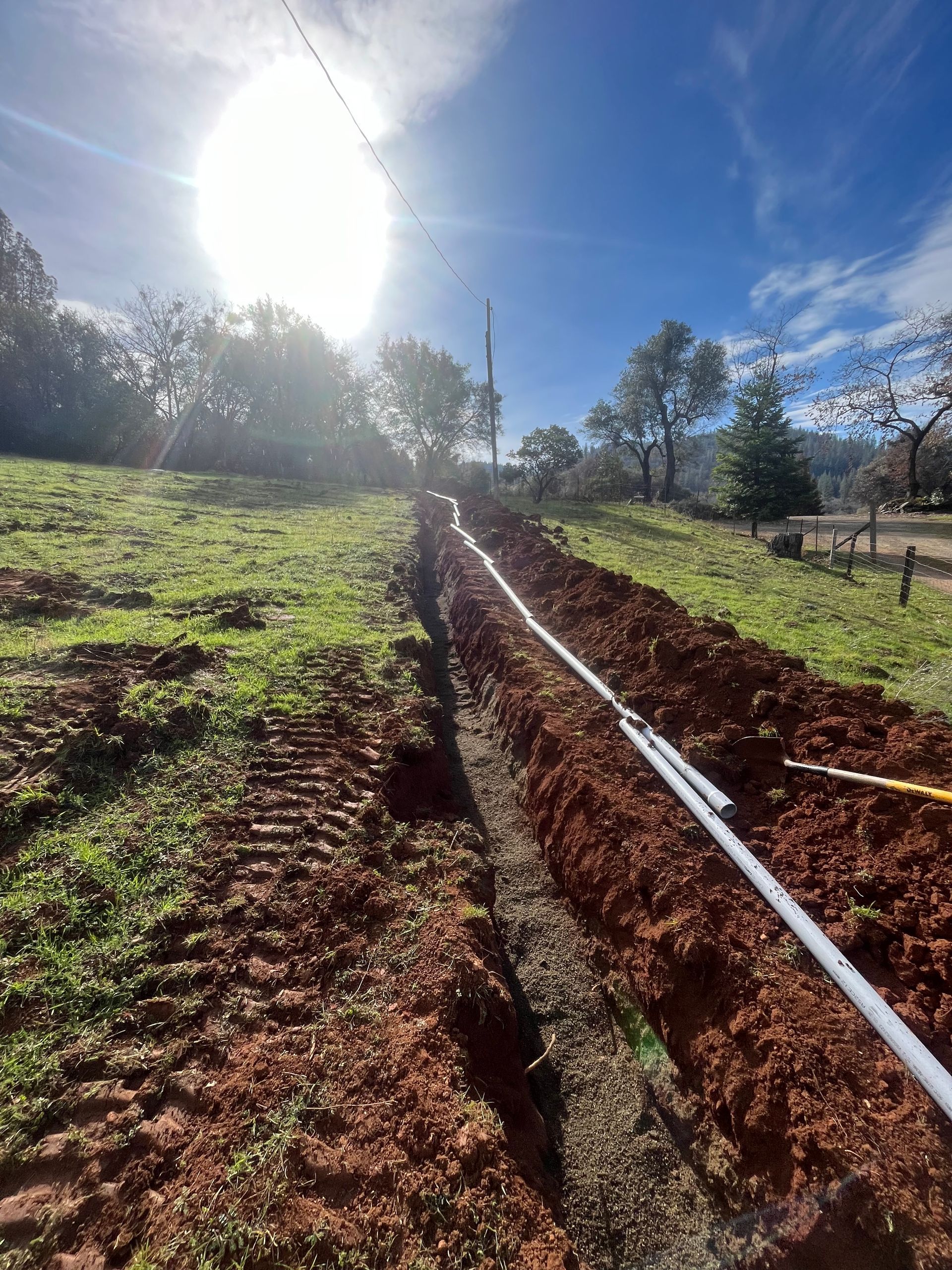 Trench in a grassy field with buried pipes, under a bright sun.