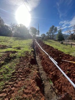 Trench in a grassy field with buried pipes, under a bright sun.