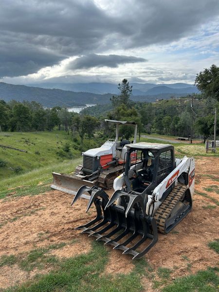 Two orange machines clearing a dirt hillside, kicking up dust under a blue sky.