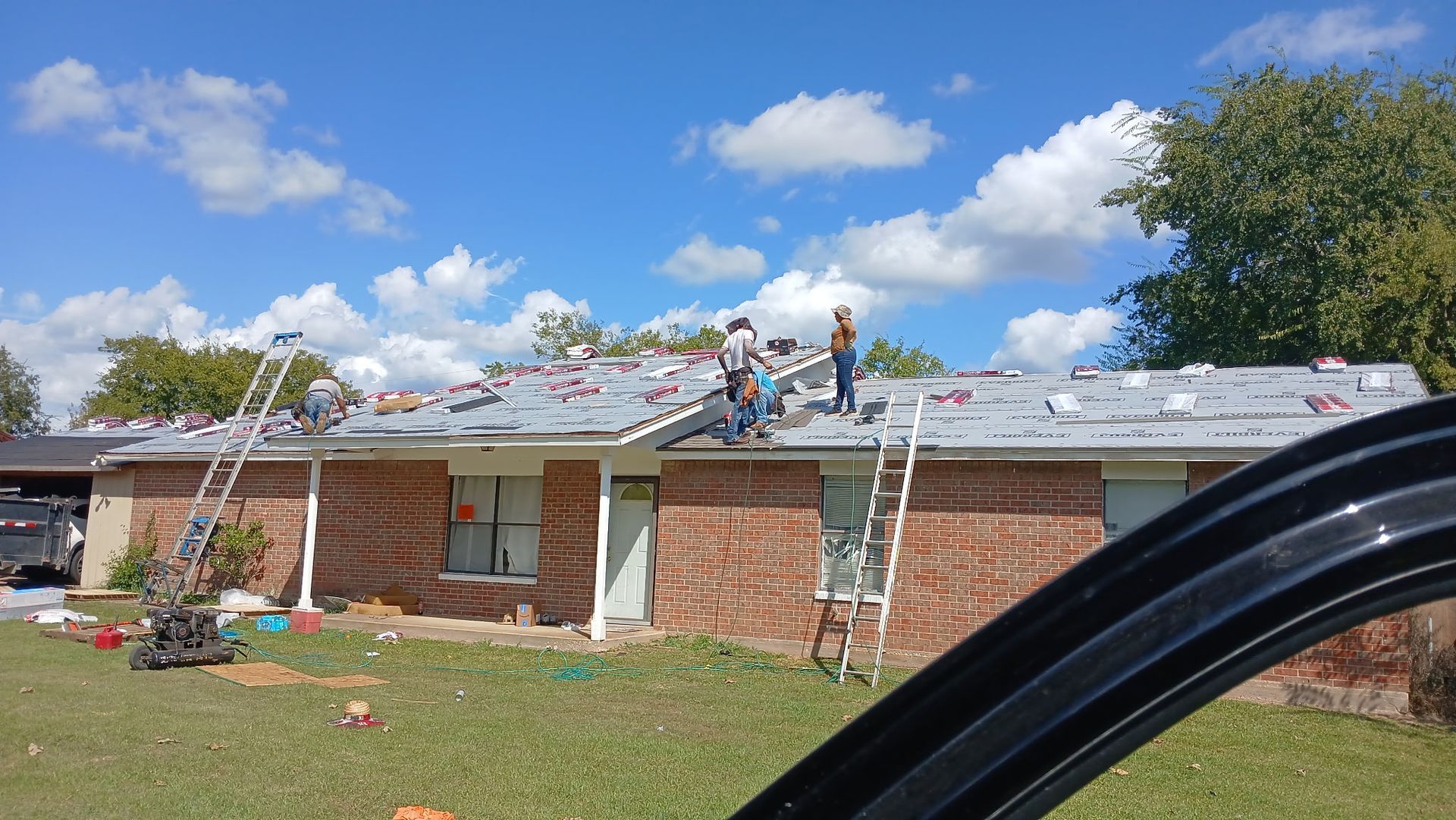 Roofers working on a one-story brick house under a blue sky with clouds. Ladders and materials visible.