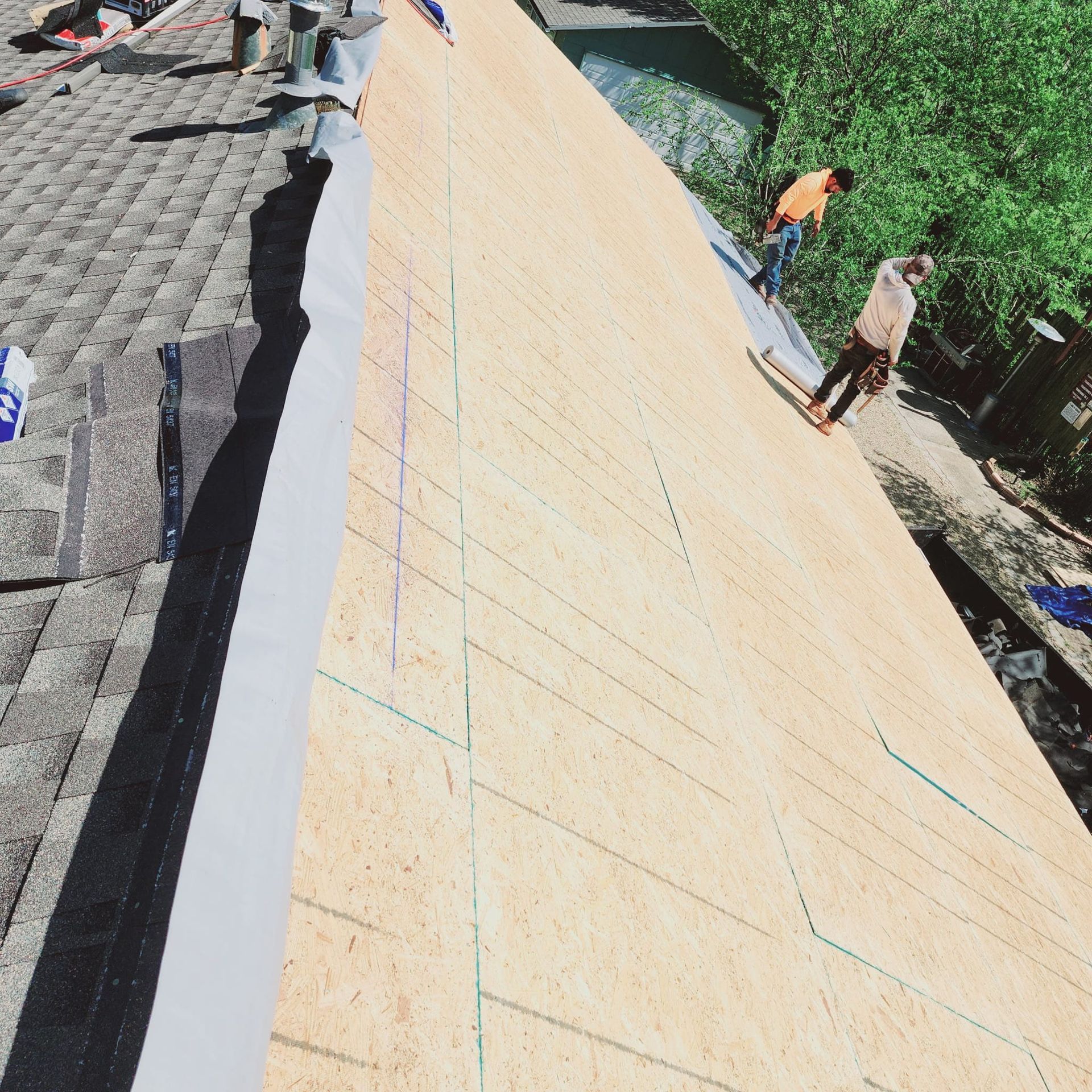 Roofers installing shingles on a house roof; two workers on the roof, one with orange shirt.