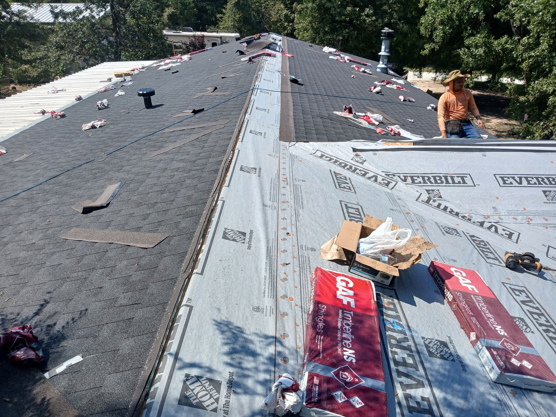 Roof partially covered in black asphalt shingles and protective underlayment; worker on roof.