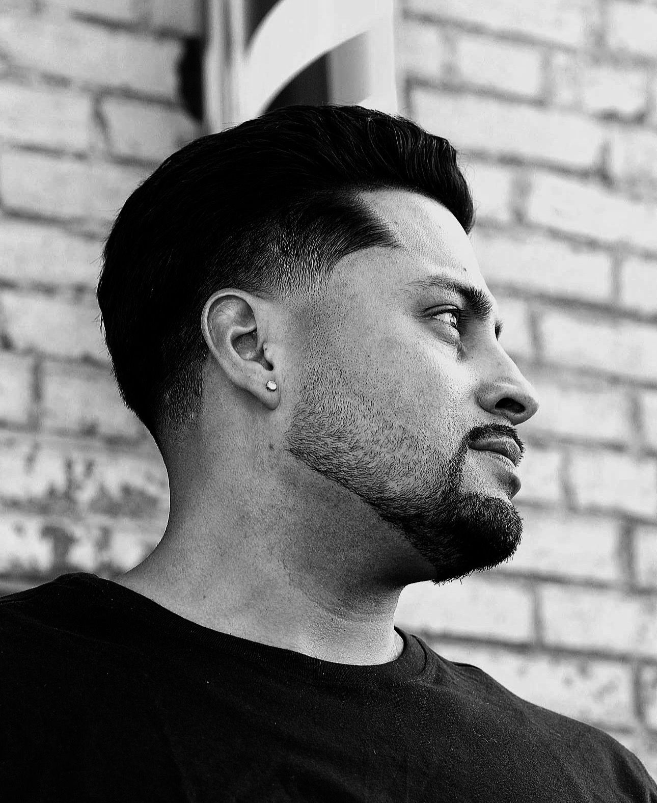 Man with short, neatly styled hair and beard, looking right, against a brick wall.