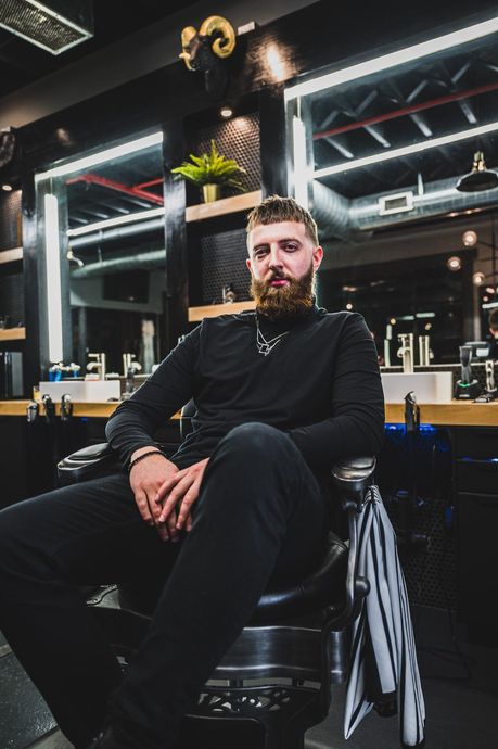 A man with a beard, in black clothes, sits in a barber chair inside a barbershop.