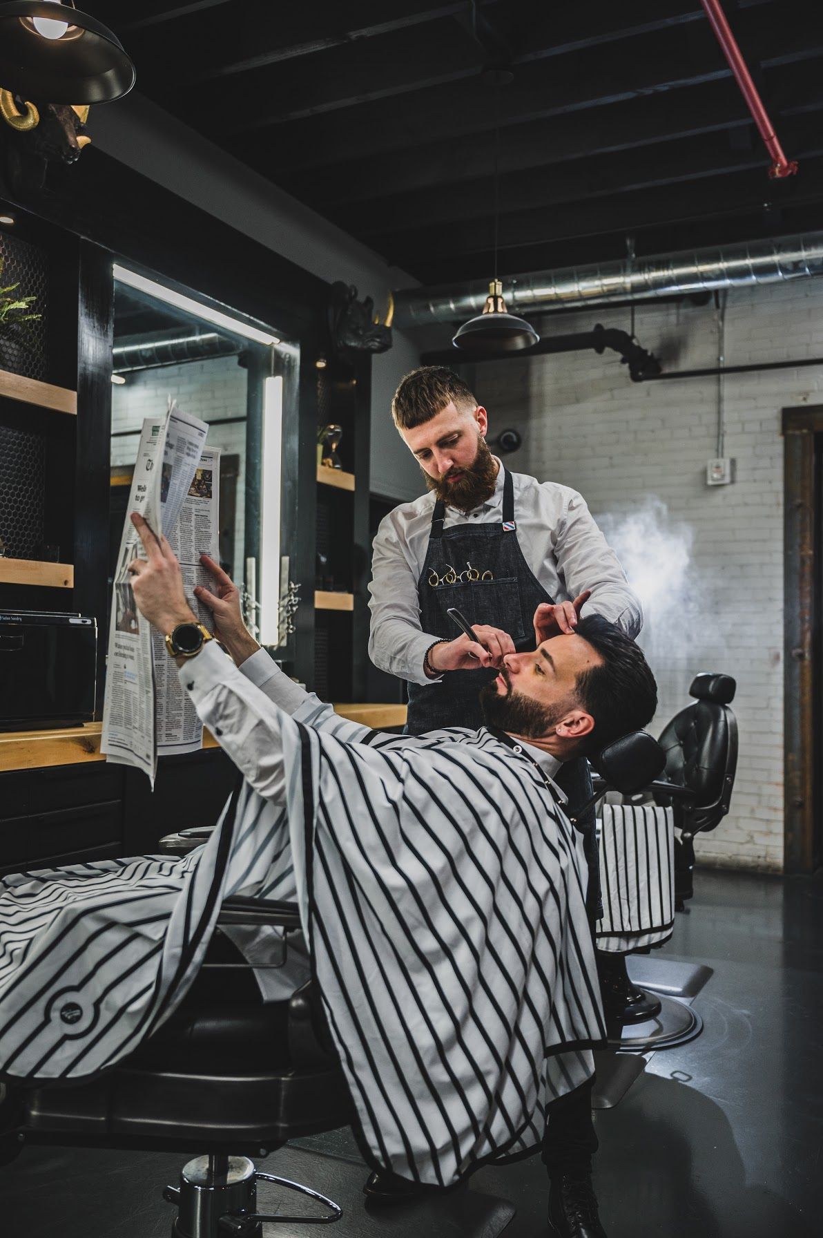 Barber trimming a man's beard in a vintage barbershop. Man is reclining, reading newspaper. Steam rises.