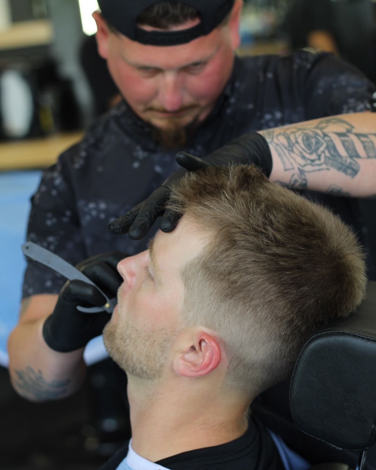 Barber shaving a client's face with a straight razor. Man in barber chair, black gloves, dark clothing.