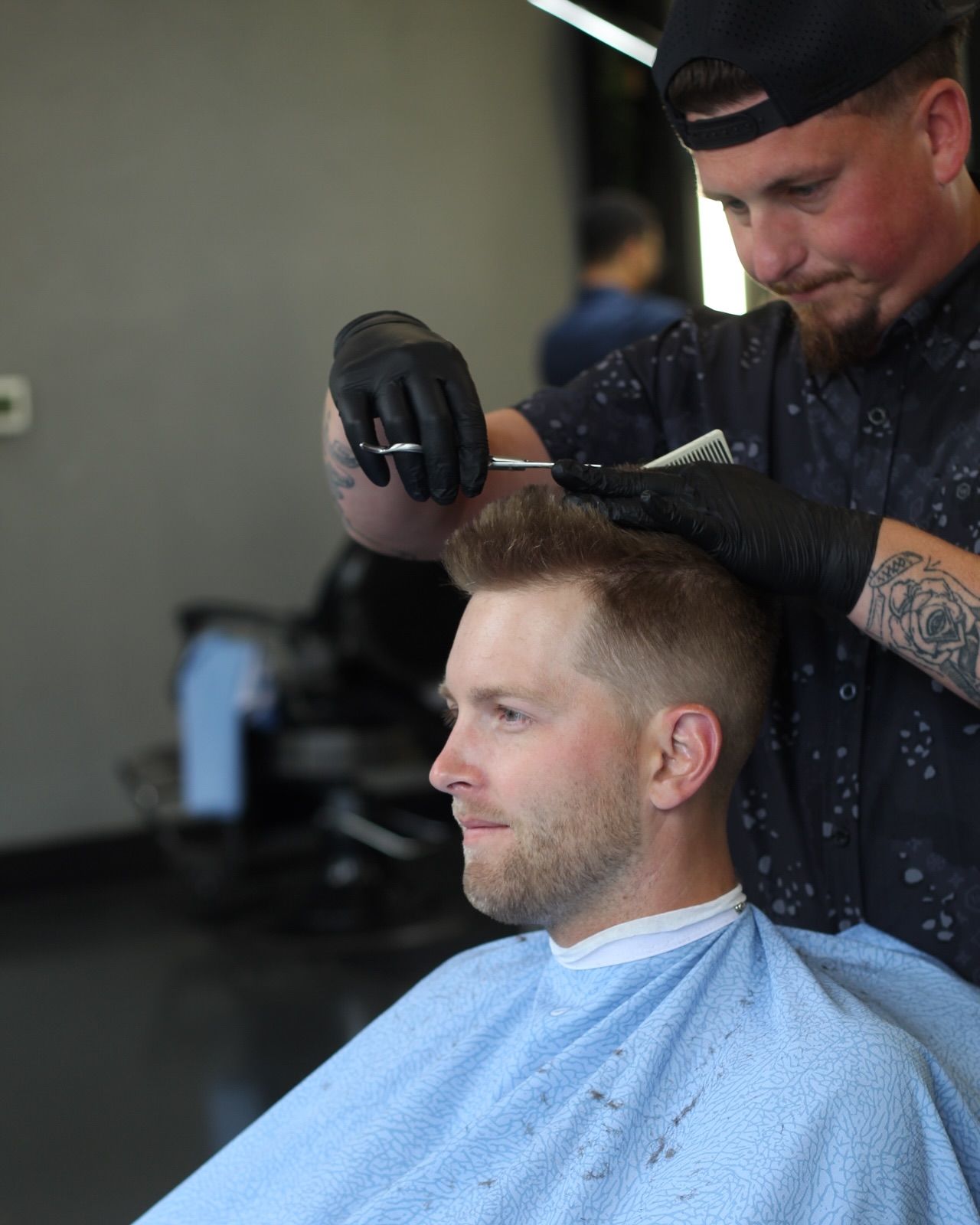Barber cutting a man's hair; man in a blue cape, barber wearing black gloves, modern salon setting.