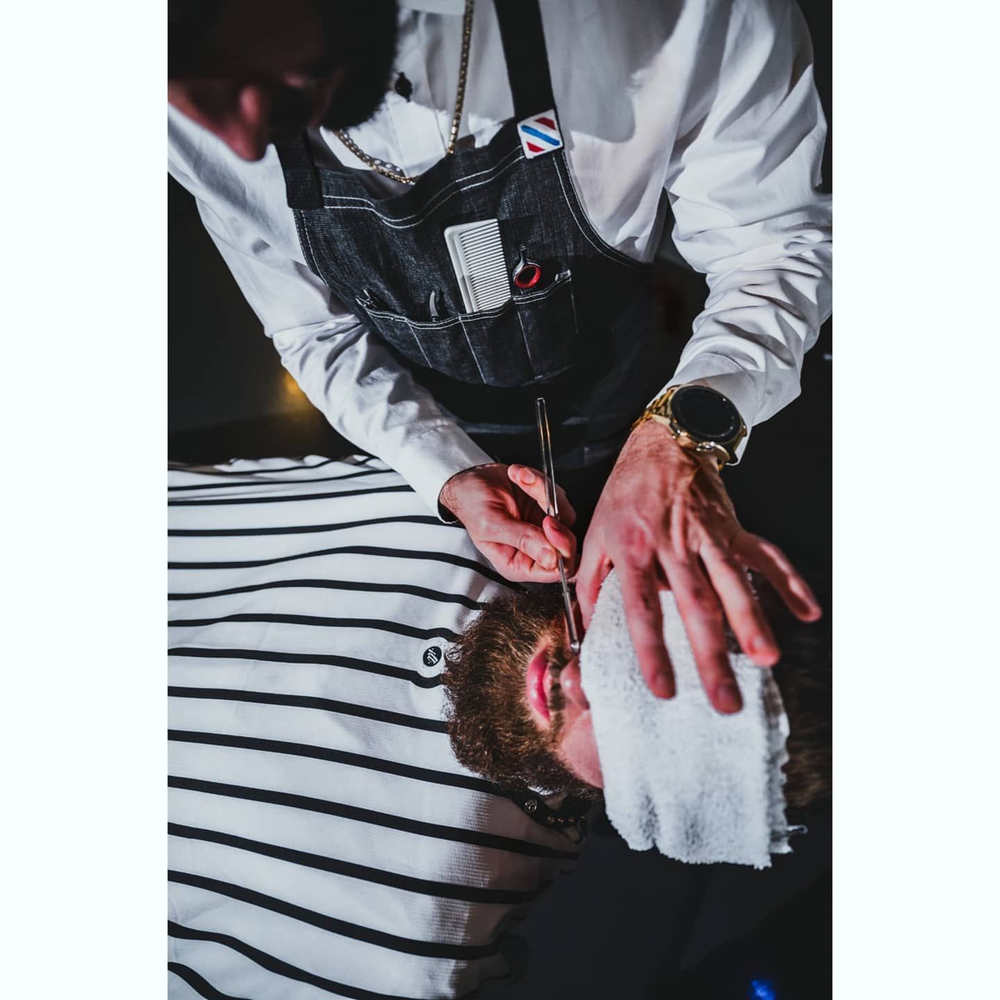 Barber shaves a client's face with a straight razor. Client has towel on face.