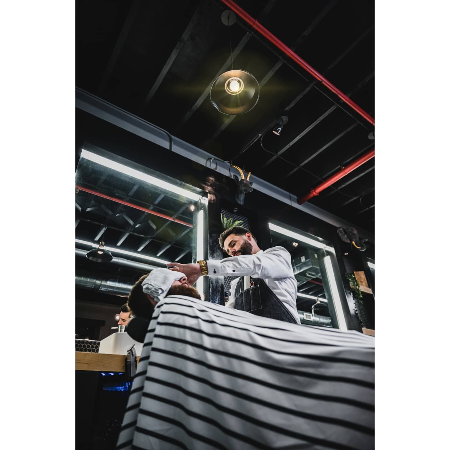 Barber shaving a customer's face in a barbershop, under a bright light. Black and white striped cape.