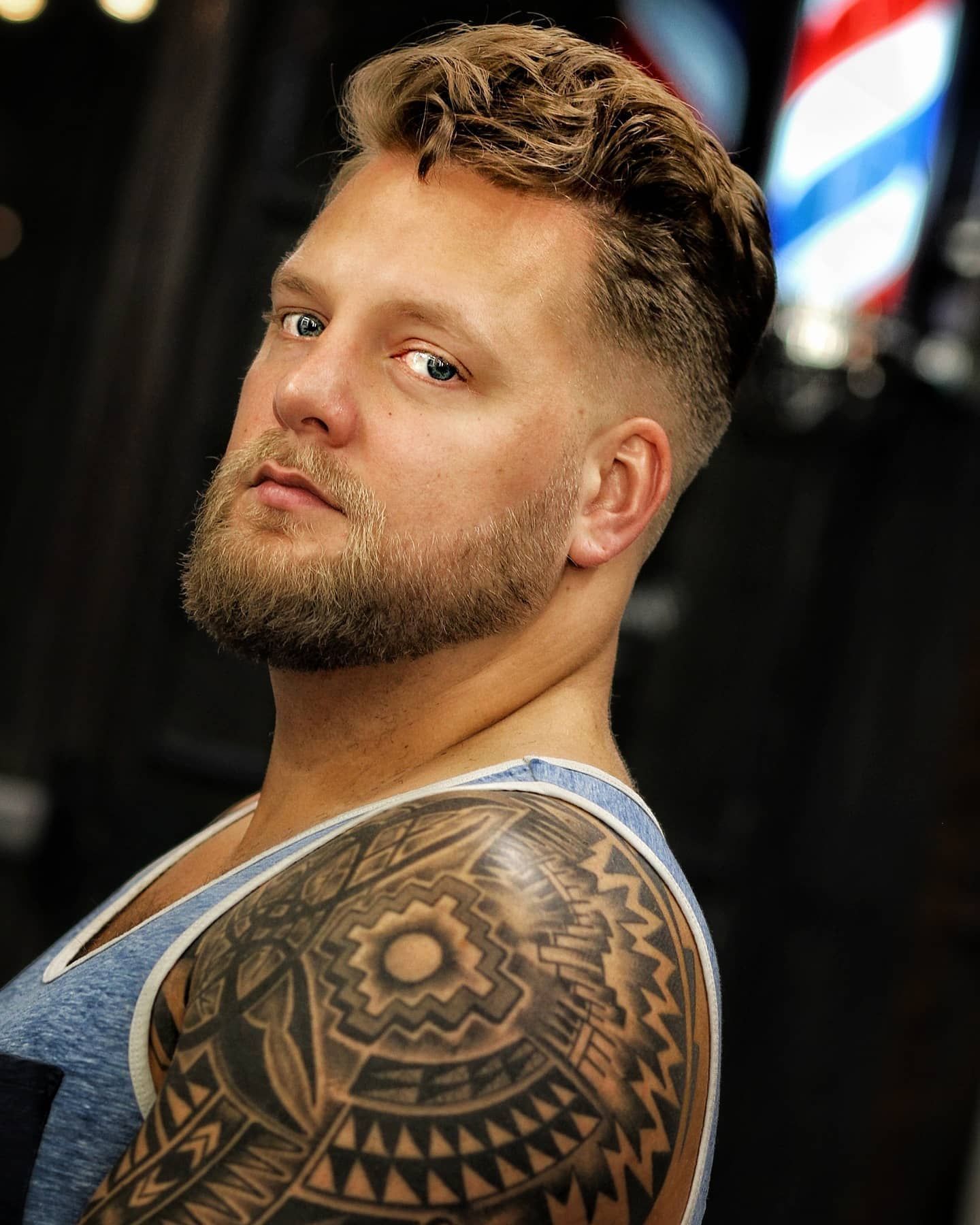 Man with a beard and tattoo, wearing a tank top in a barbershop.