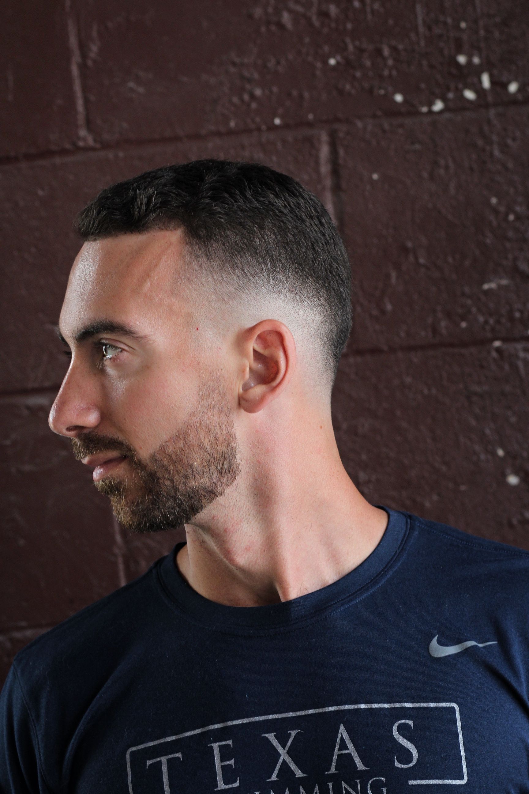 Man with fade haircut and beard, wearing a navy blue shirt, standing against a brick wall.