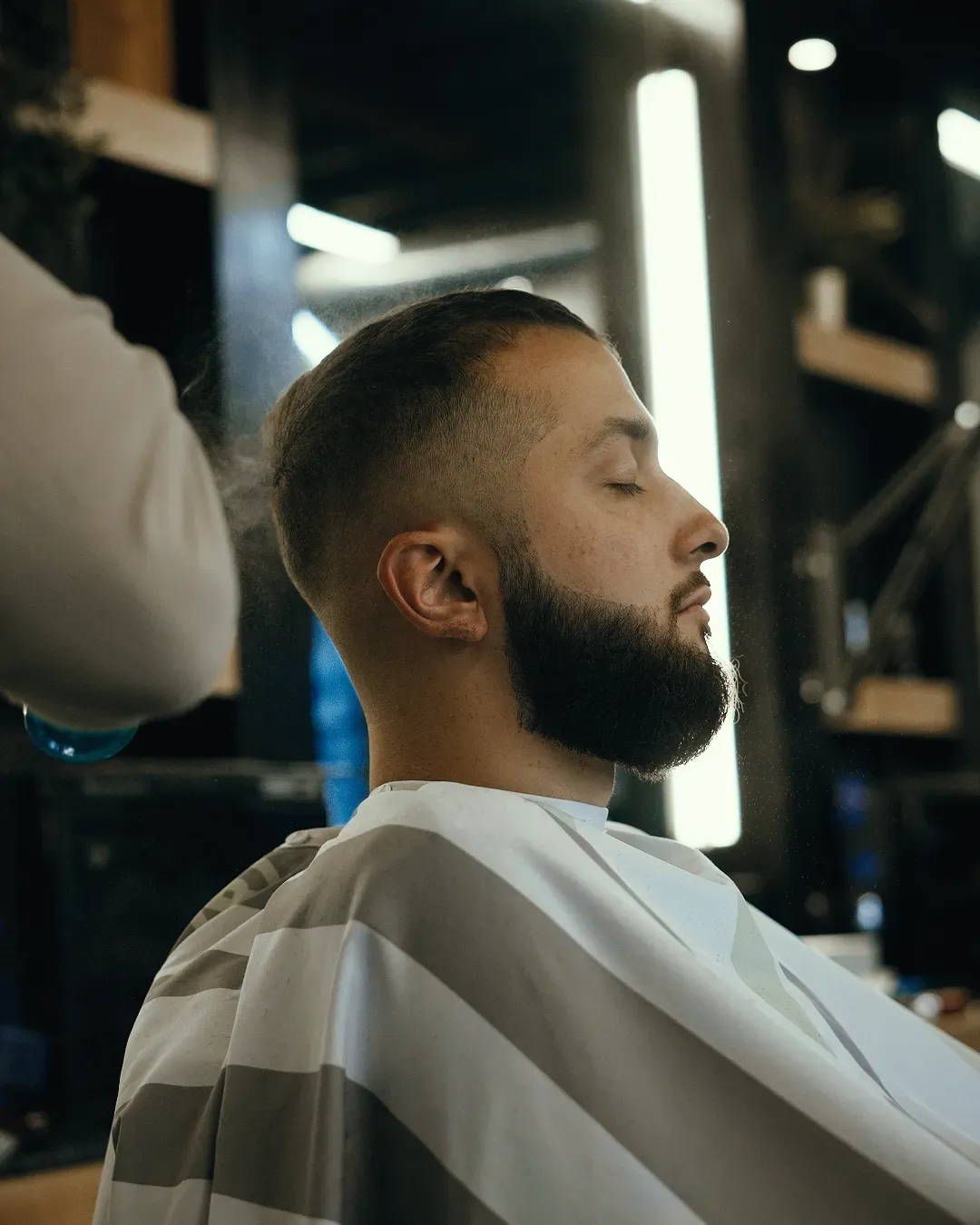 Man getting a haircut; barber shop setting. Side profile of man with beard, eyes closed, wearing barber cape.