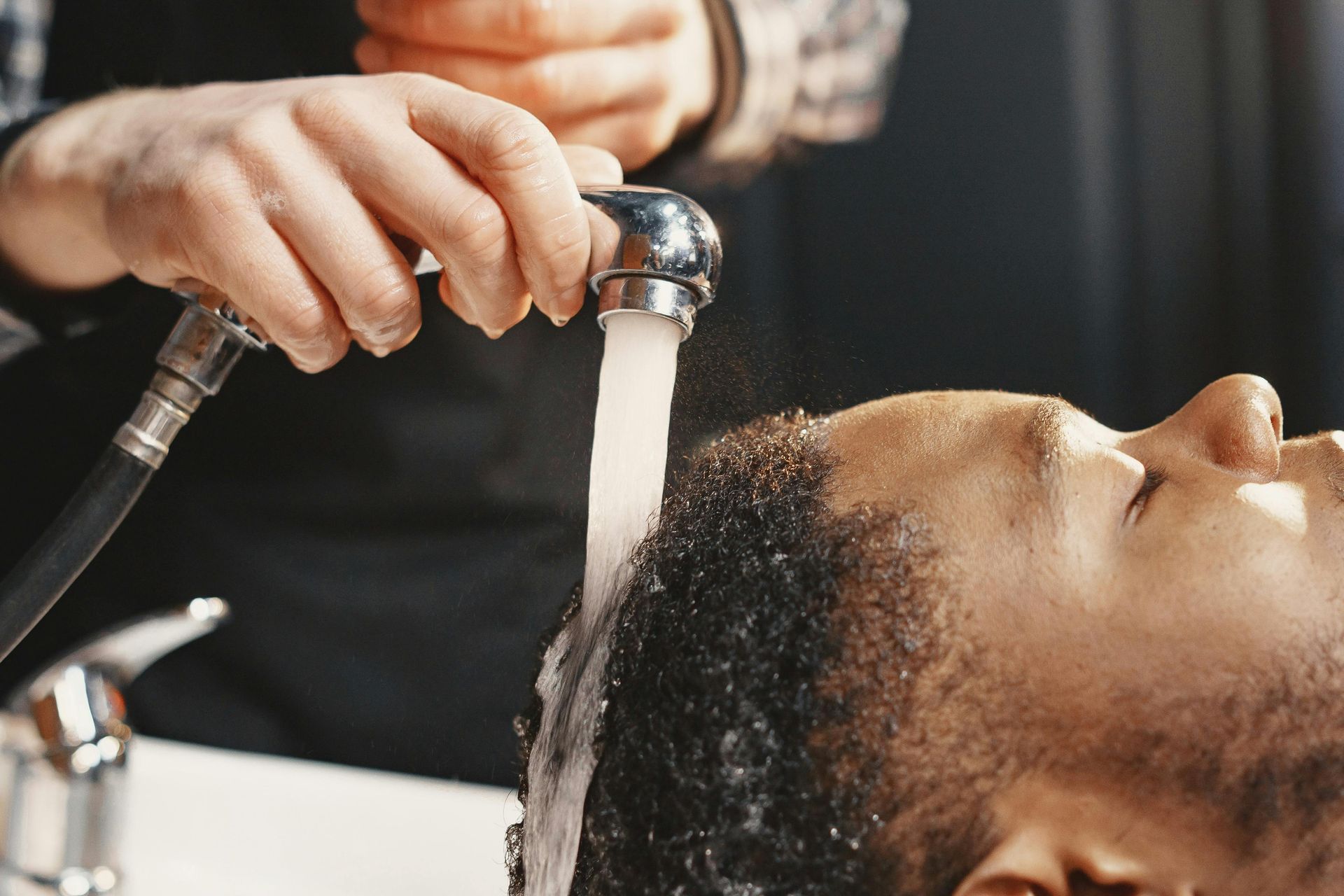 Barber washing a man's hair in a salon. The man is relaxed with his eyes closed as the barber pours water over his head.