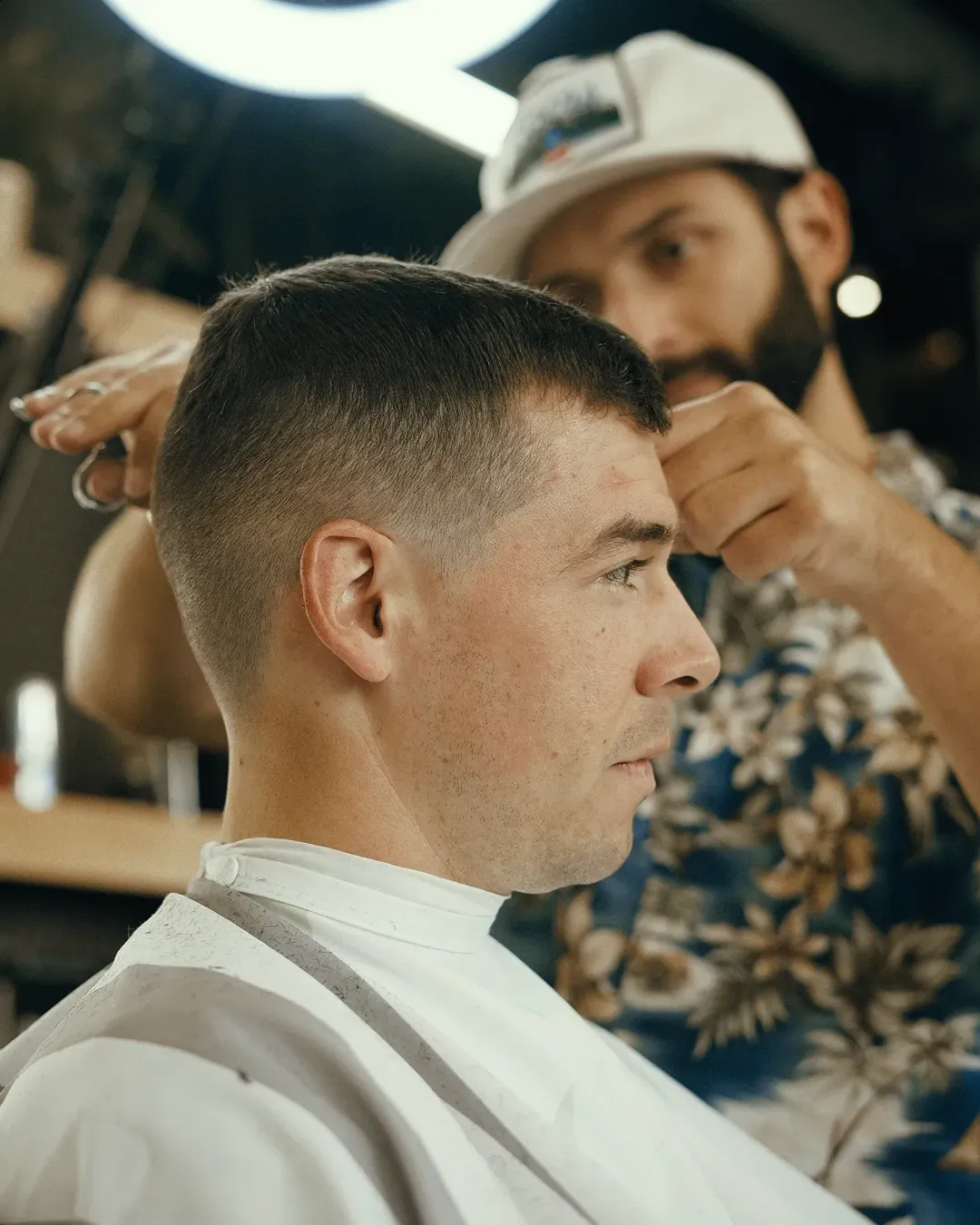 A barber cutting a man's short hair in a salon; the barber is wearing a hat and patterned shirt.