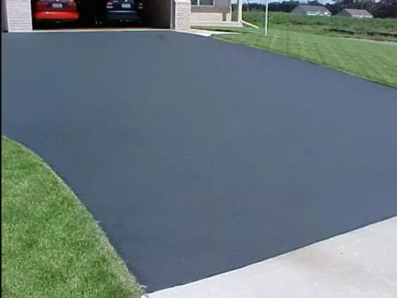 A newly paved dark gray asphalt driveway with a garage in the background, bordered by green grass.