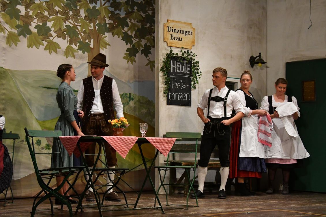 Fünf Schauspieler der Theatergruppe Emmering auf der Bühne vor einer Wirtshauskulisse, zwei reden miteinander.