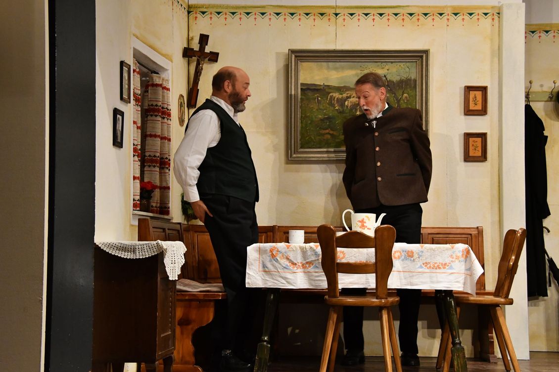 Zwei Schauspieler der Theatergruppe Emmering stehen an einem Tisch auf der Bühne im Stück 