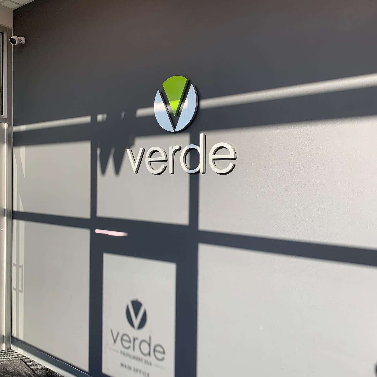 The Verde office building entrance featuring the company logo on a light wall with shadows across the glass door.