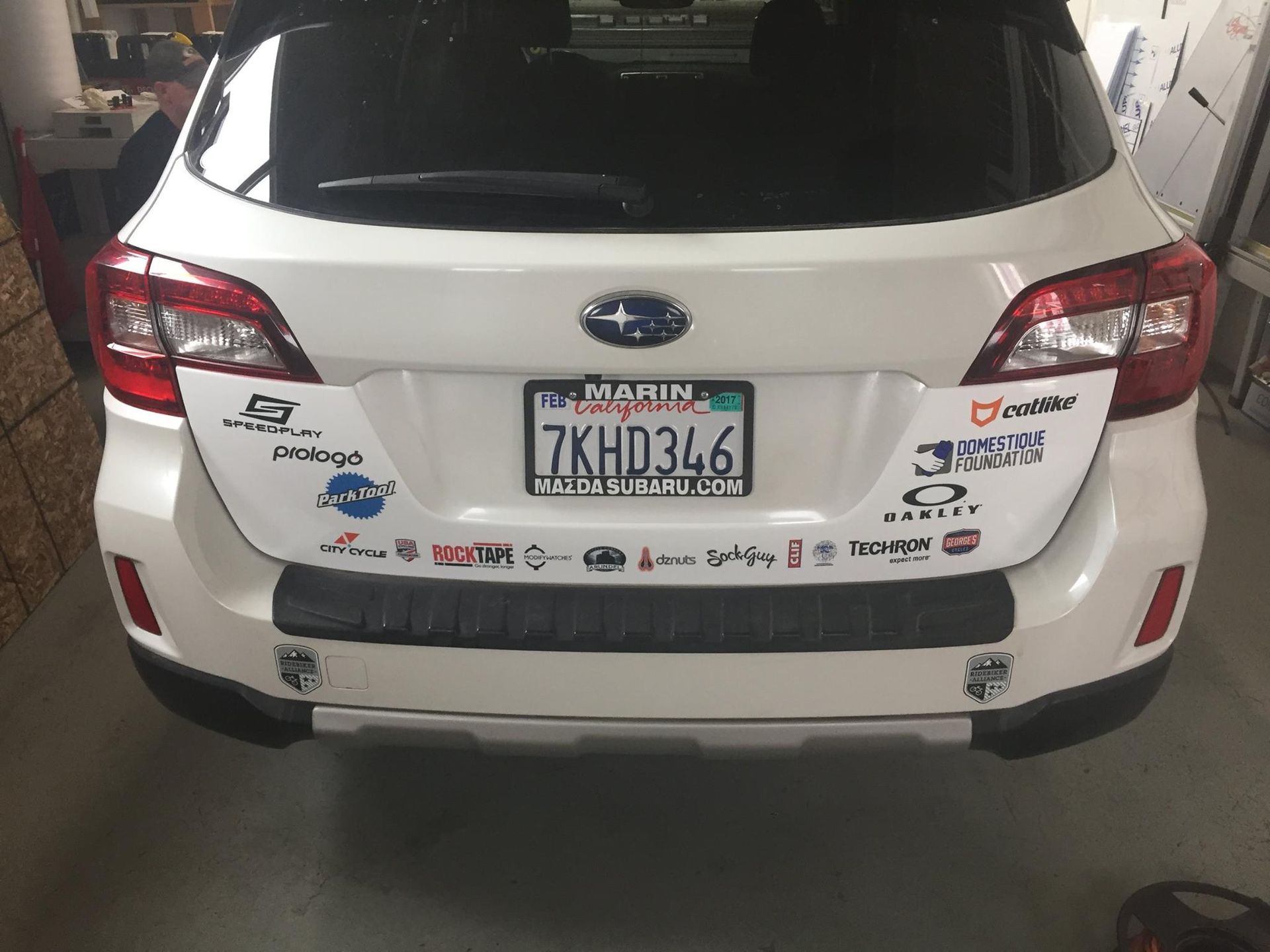 White Subaru SUV parked indoors, featuring a California license plate 7KHD346 and numerous stickers on the trunk.