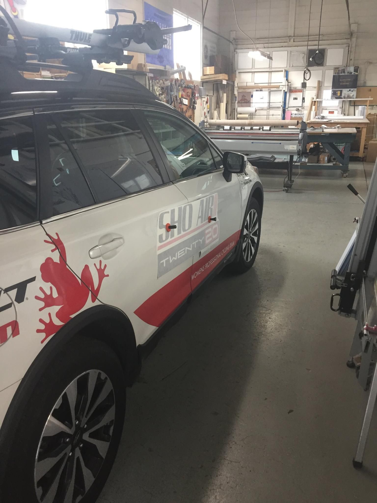 A white vehicle with red frog graphics parked inside an automotive workshop with work tables and equipment in the background.