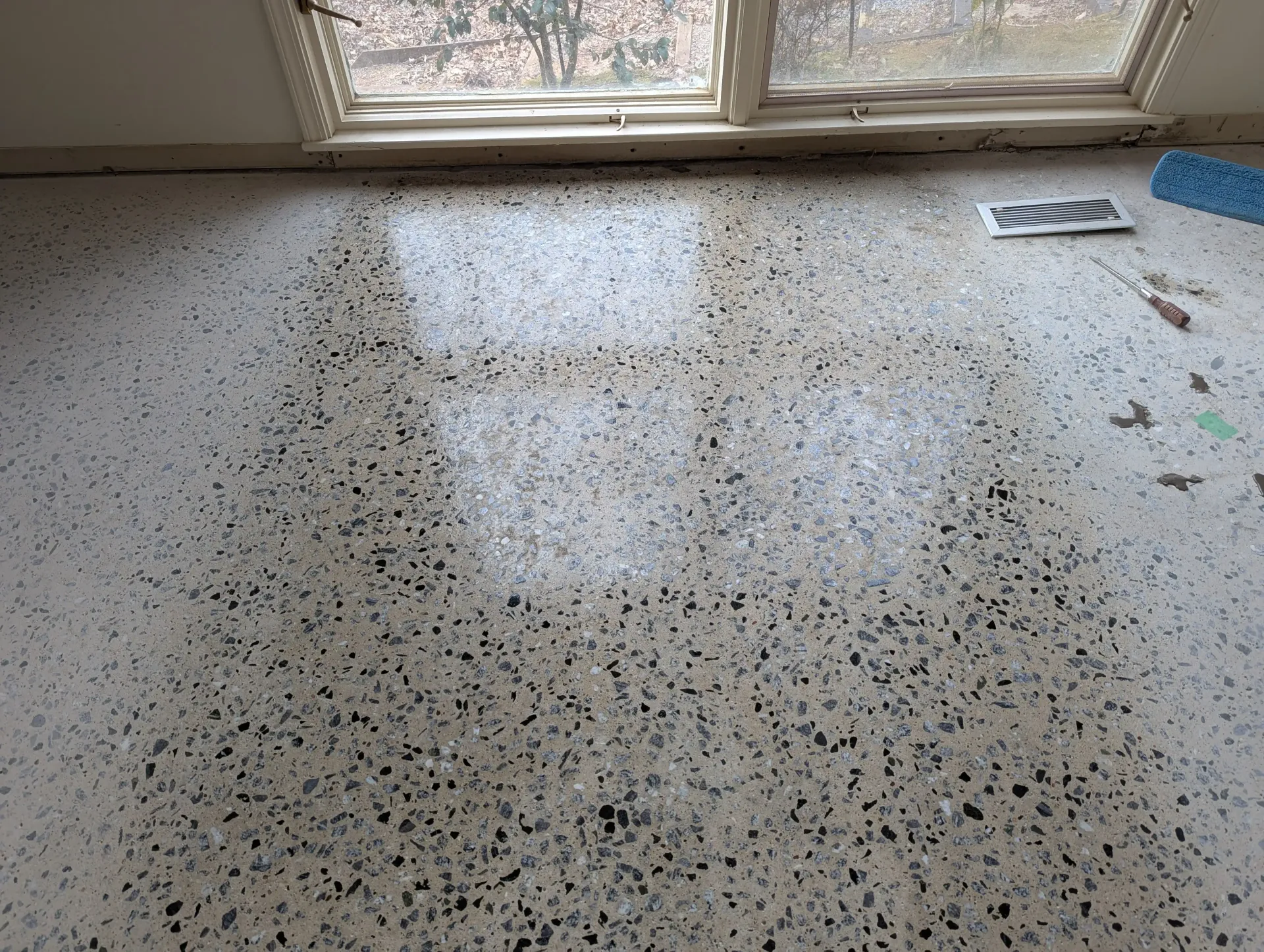 A speckled terrazzo floor showing a reflective, wet patch in front of a window, with a floor vent nearby.