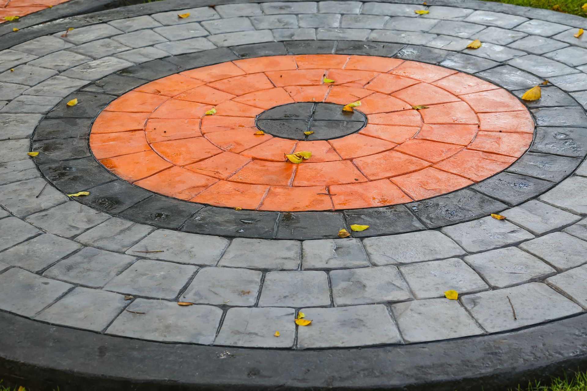 Circular stone patio with concentric rings in gray, black, and orange. Scattered yellow leaves.