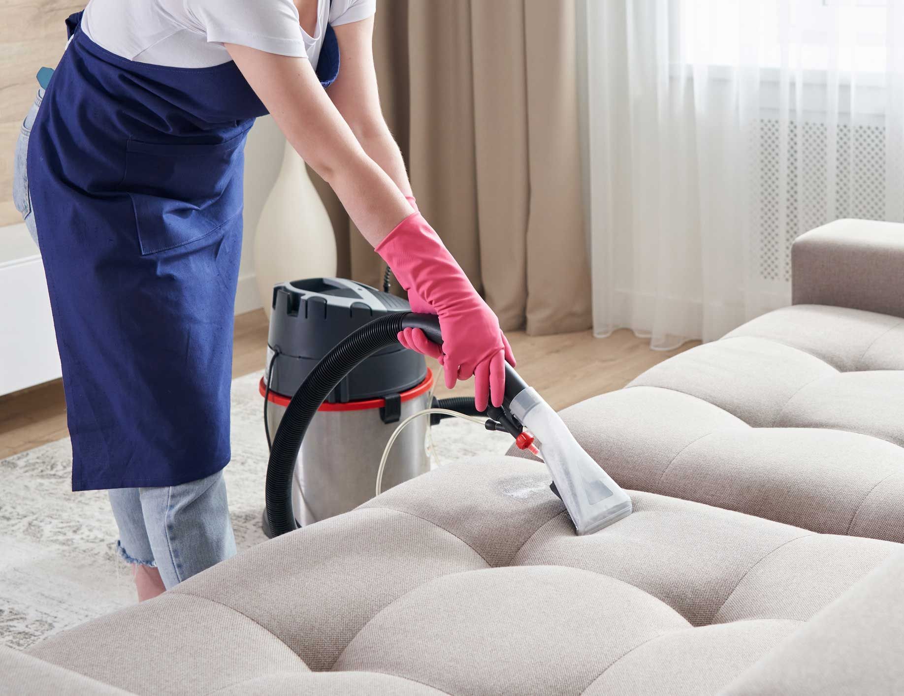 Person in pink gloves cleans a light beige couch with a vacuum in a living room.