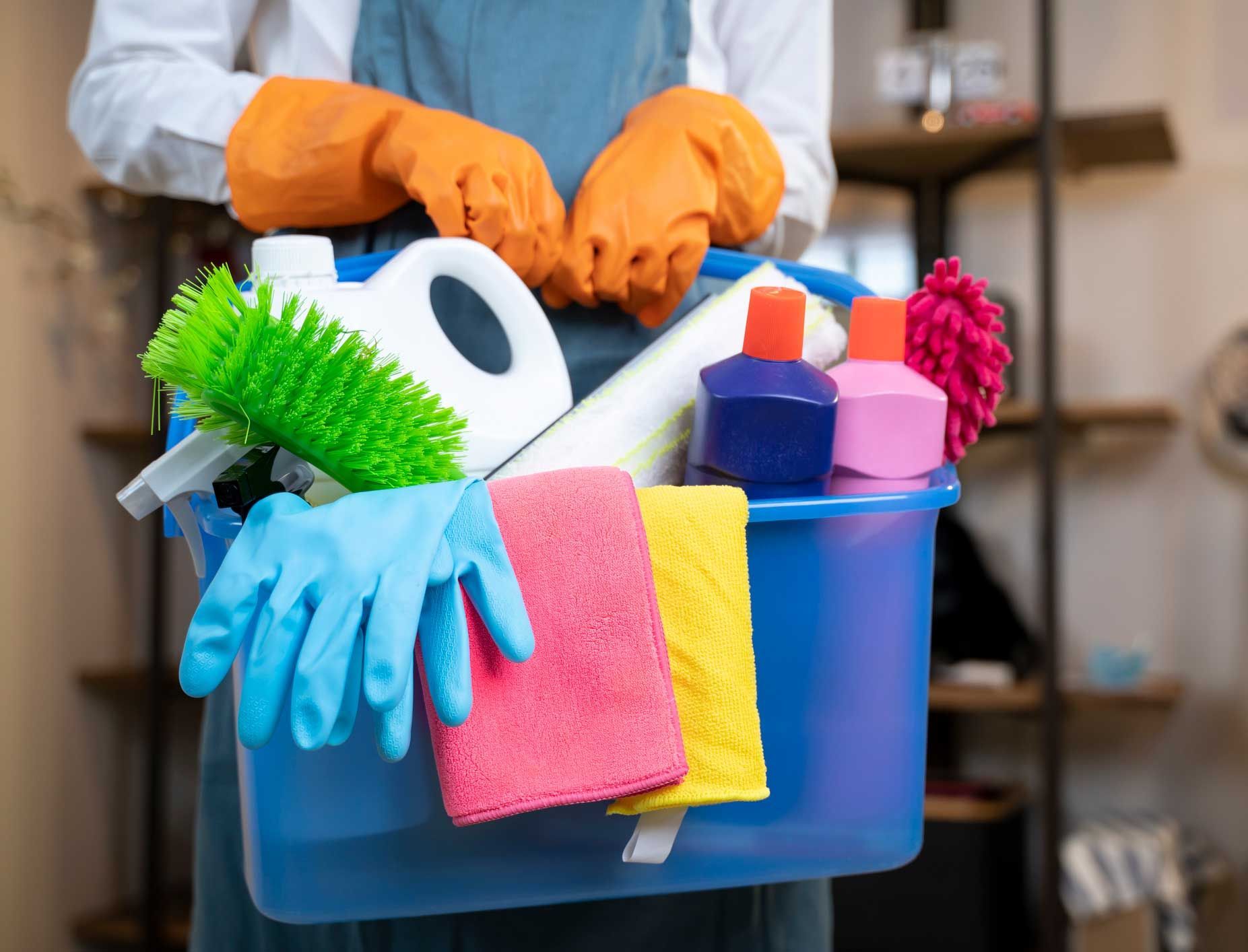 Person holding a blue bucket filled with cleaning supplies, including bottles, gloves, brushes, and cloths.