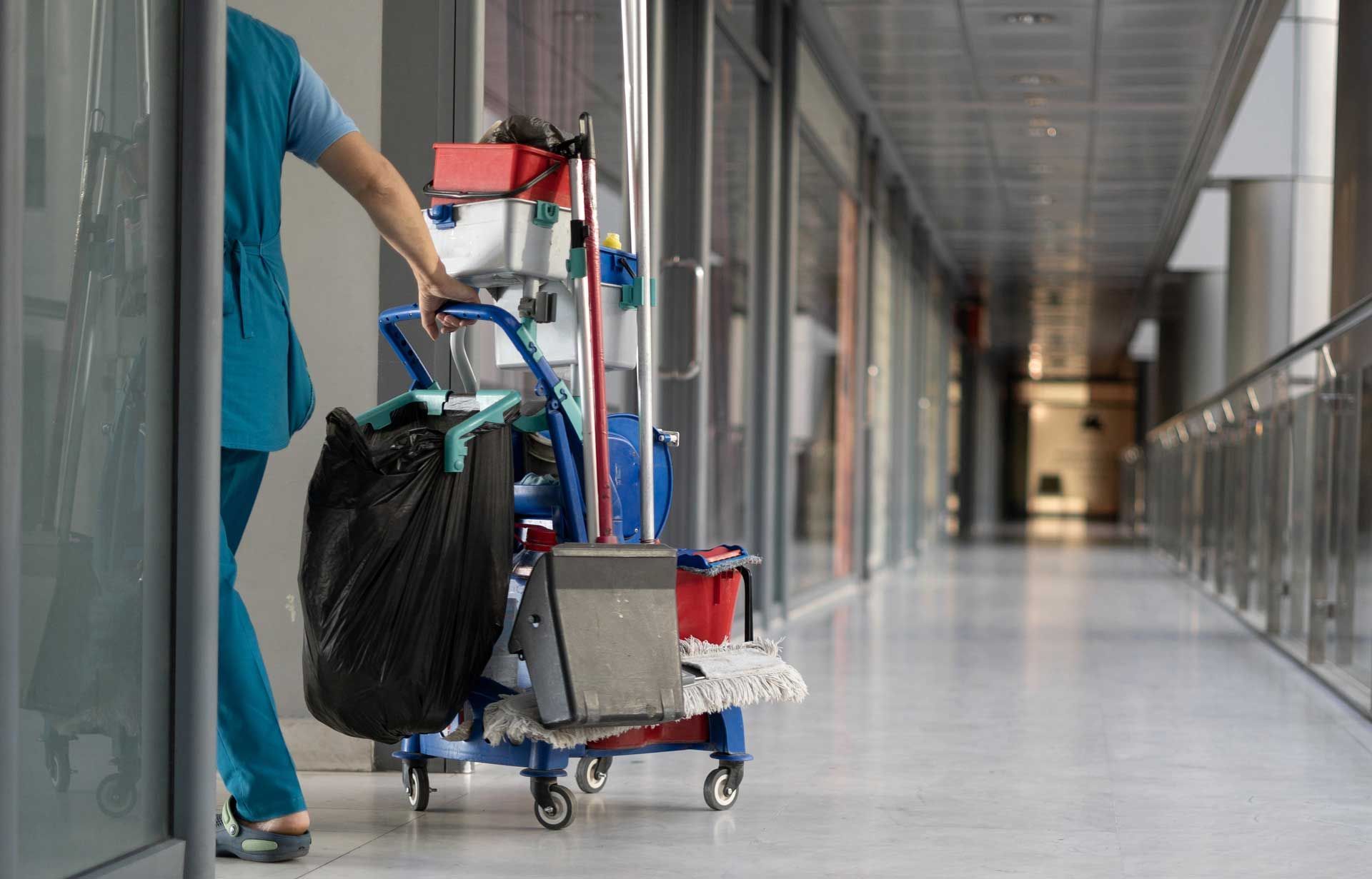 Custodian pushing a cleaning cart down a hallway with glass windows.