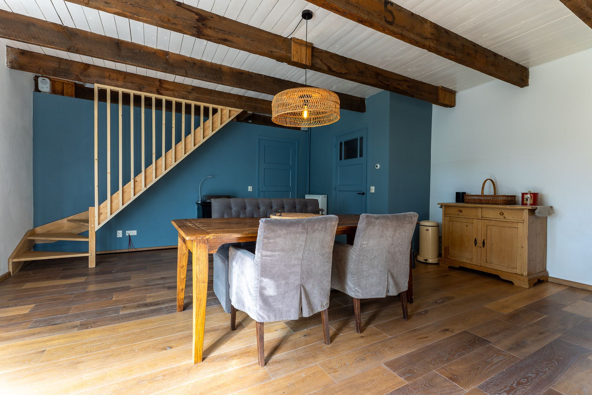 Eetkamer met houten balken, tafel en stoelen, trap en blauwe muren.