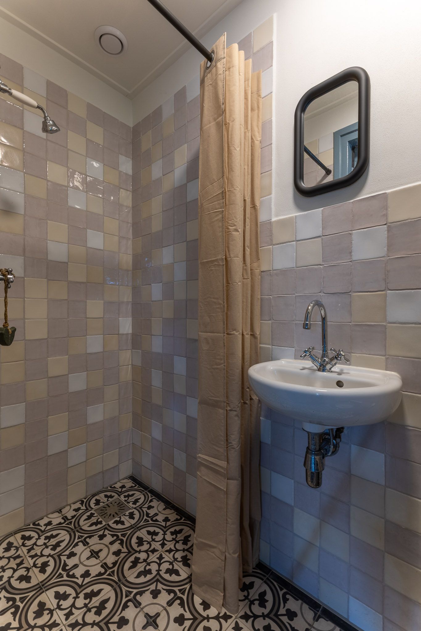 Kleine badkamer met tegelvloer met patroon, vierkante wandtegels, douchegordijn, wastafel en spiegel.