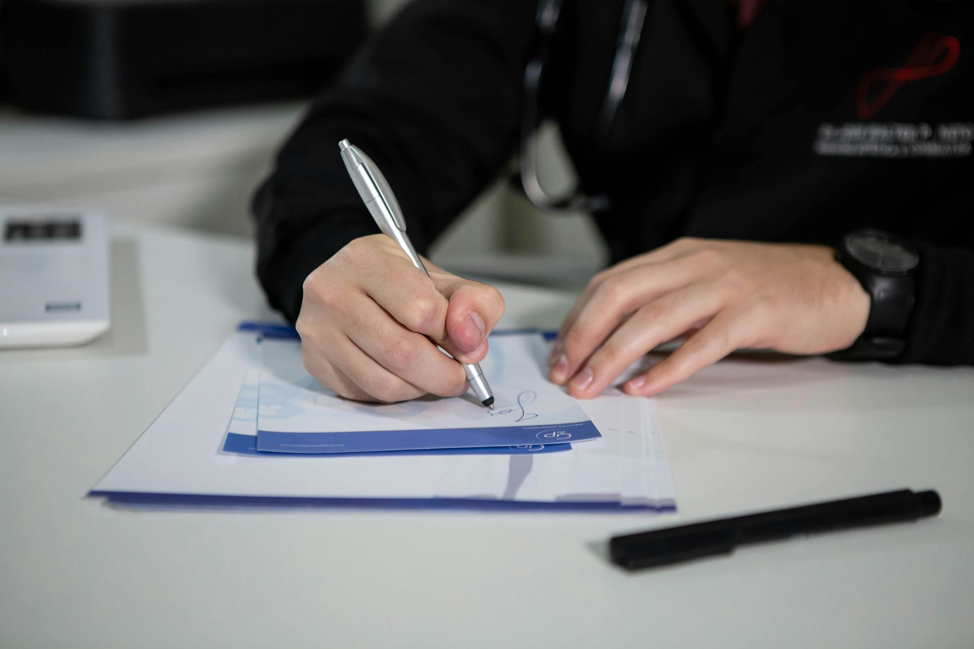Person in a black coat writing on a prescription pad at a desk; a stethoscope and watch are visible.