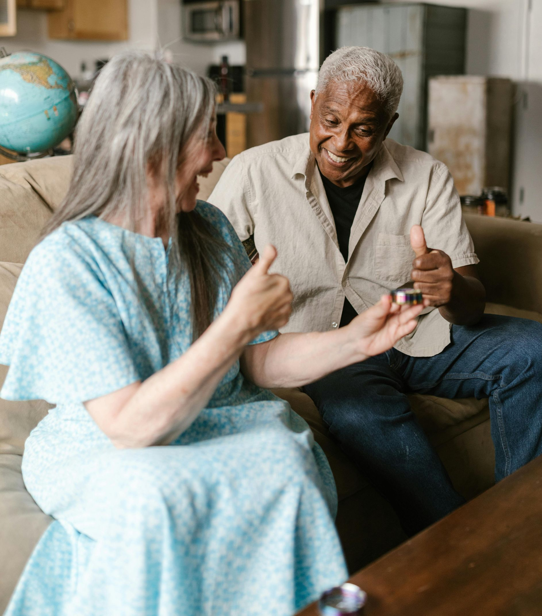 Smiling people on couch, one holding small object, both giving thumbs up.