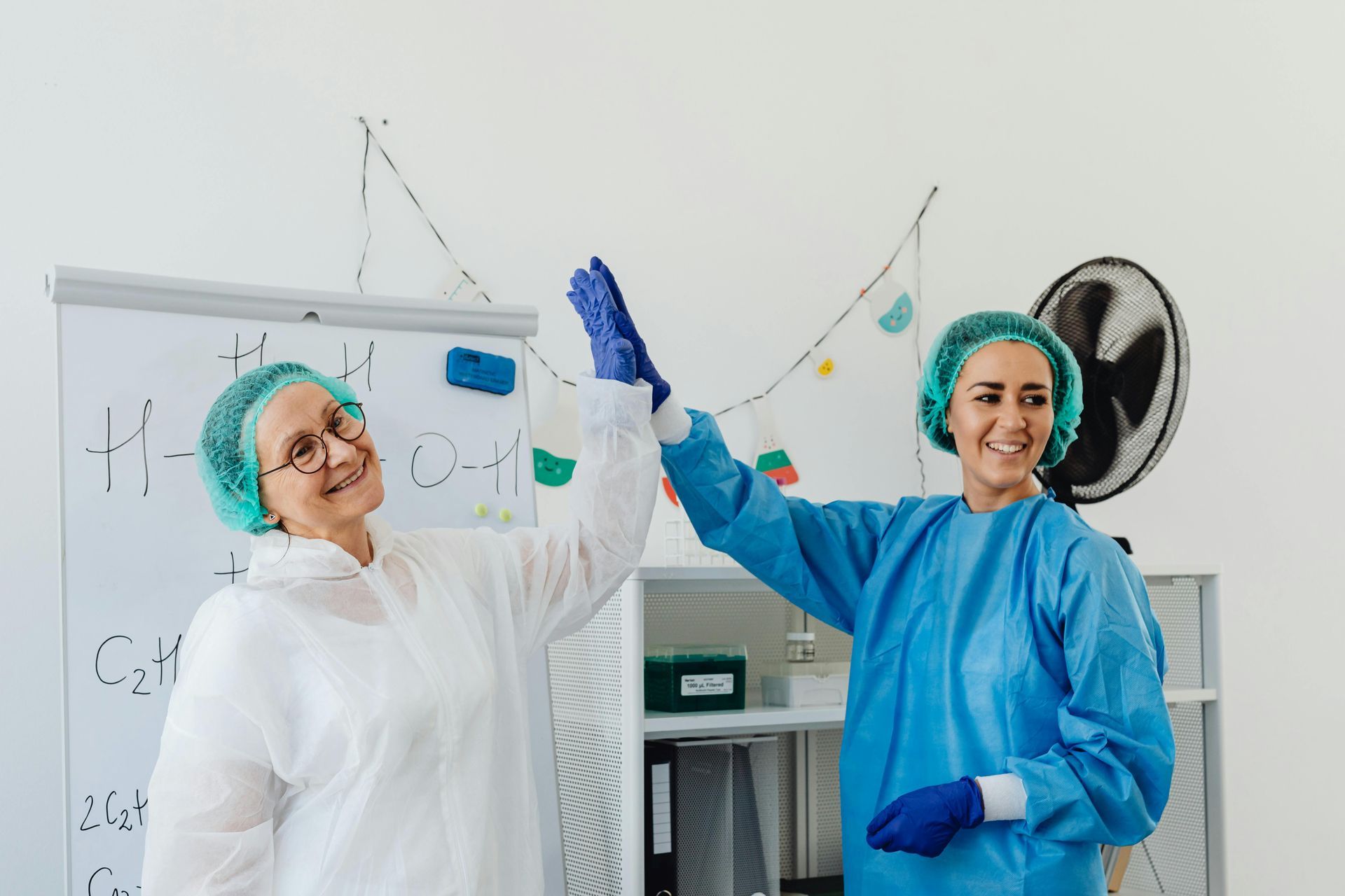 Two medical professionals in scrubs give each other a high-five, smiling. They're in a brightly lit room.