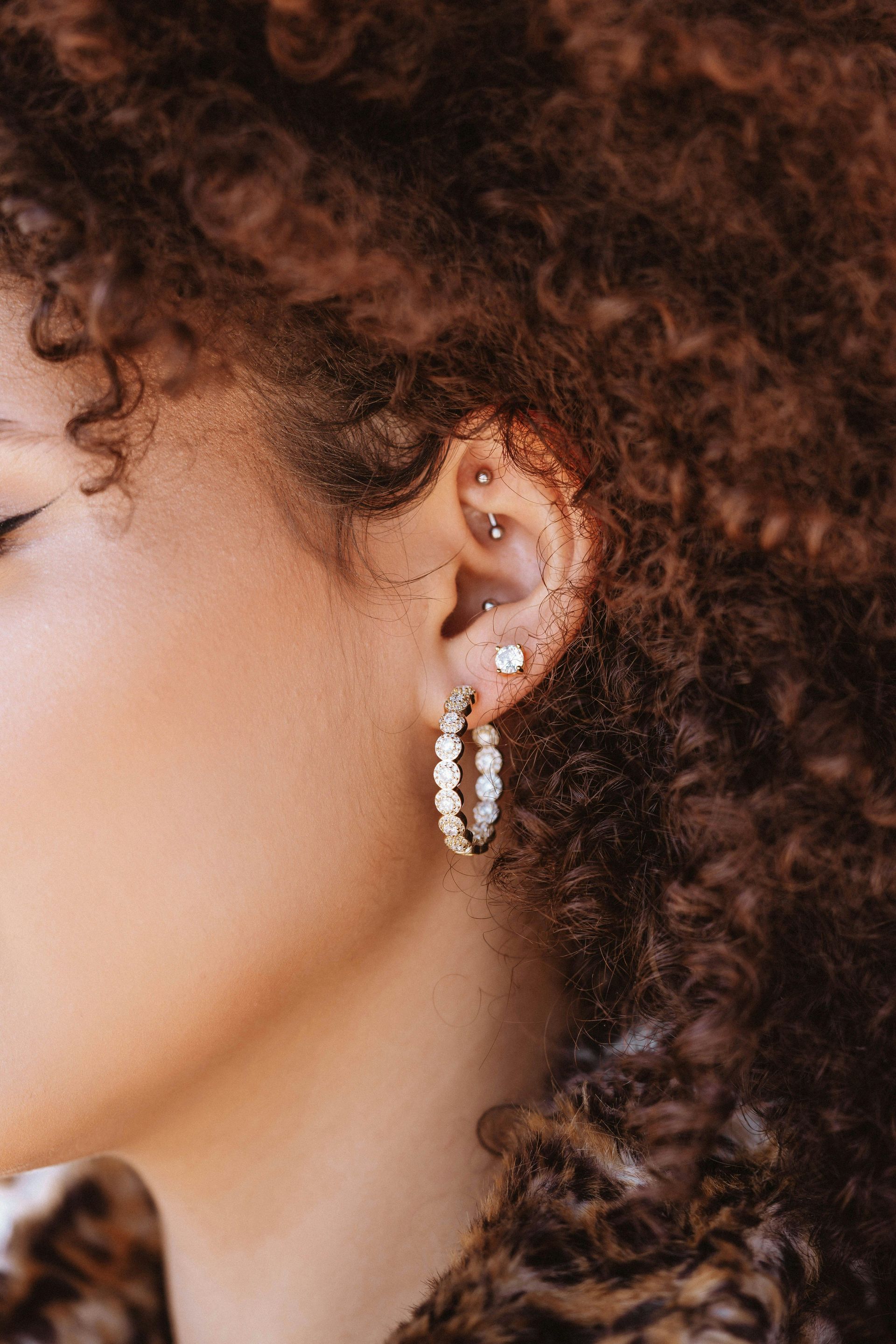 Close-up of a person's ear with hoop earrings and multiple piercings in curly brown hair.
