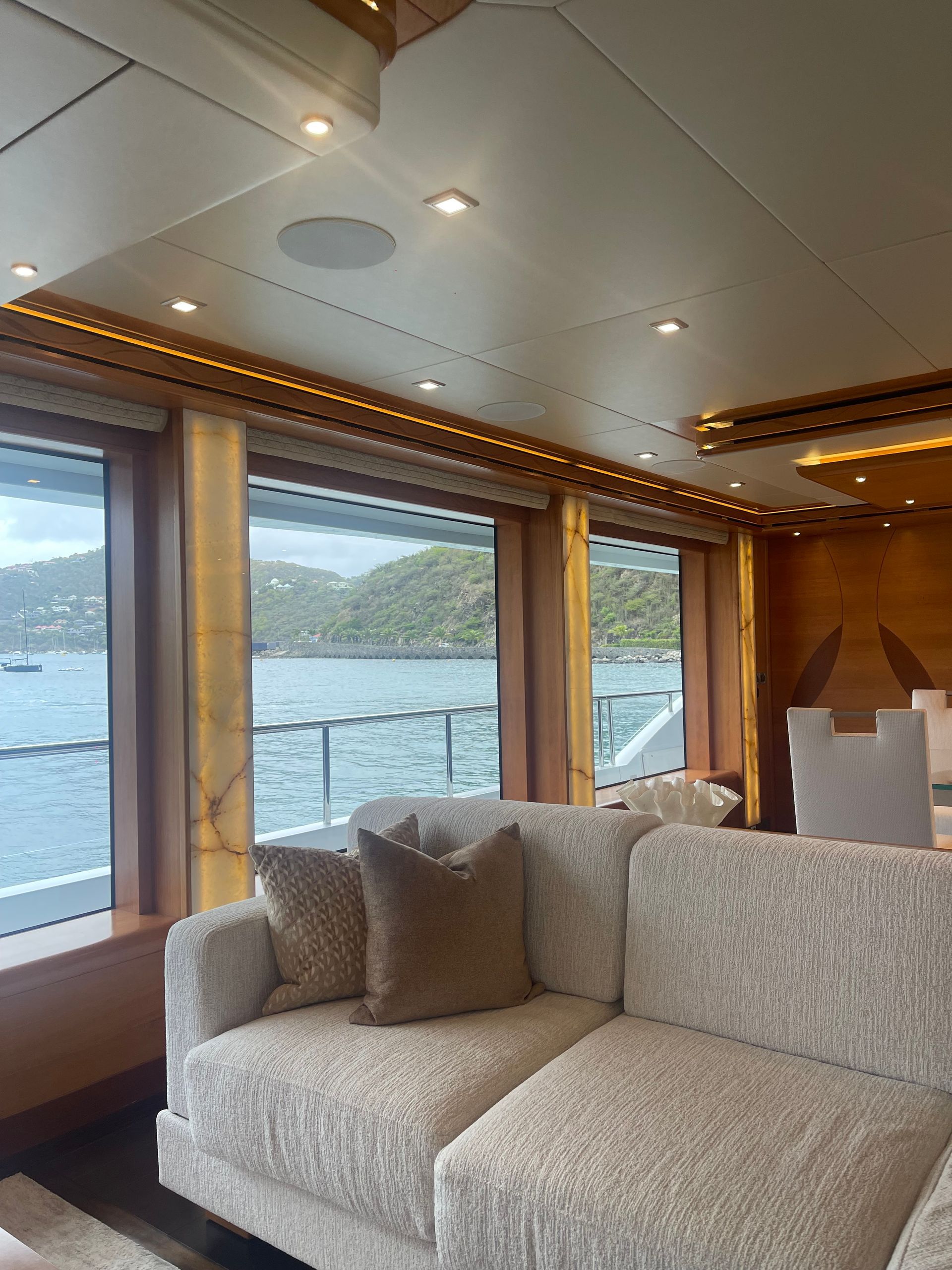 Interior of a yacht cabin with a sofa, large windows overlooking water and a mountain, and built-in lighting.