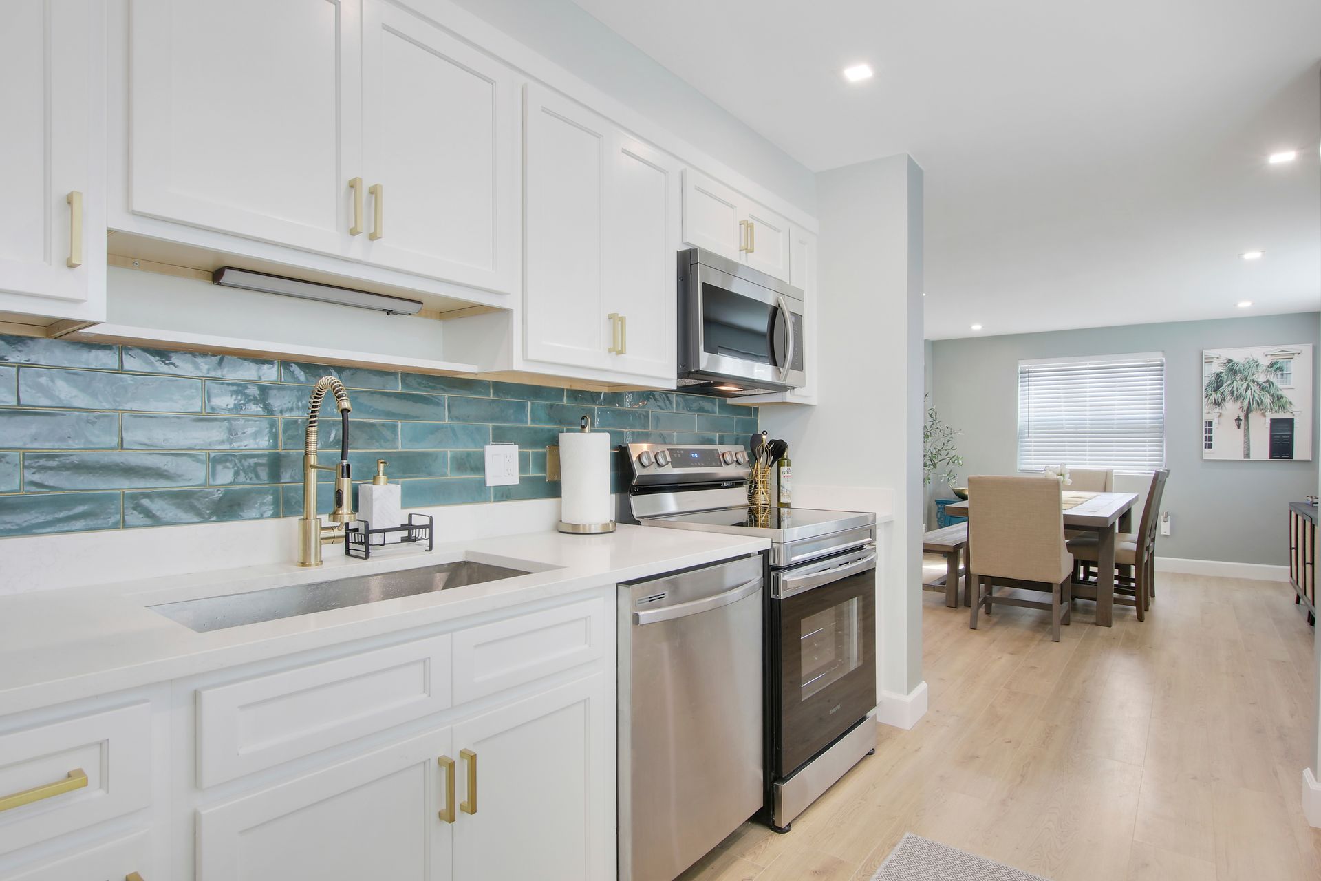 Bright kitchen with white cabinets, stainless steel appliances, and teal backsplash, leading to a dining room.