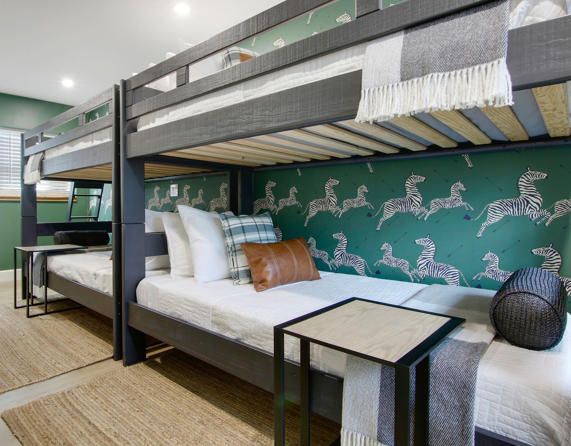 Two dark gray bunk beds in a room with green and zebra-print wallpaper, beige rugs, and side tables.