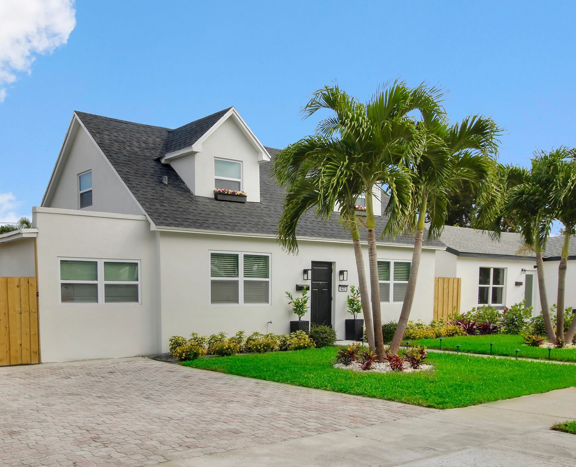 White house with black roof, palm trees, and green lawn. Sunny day.