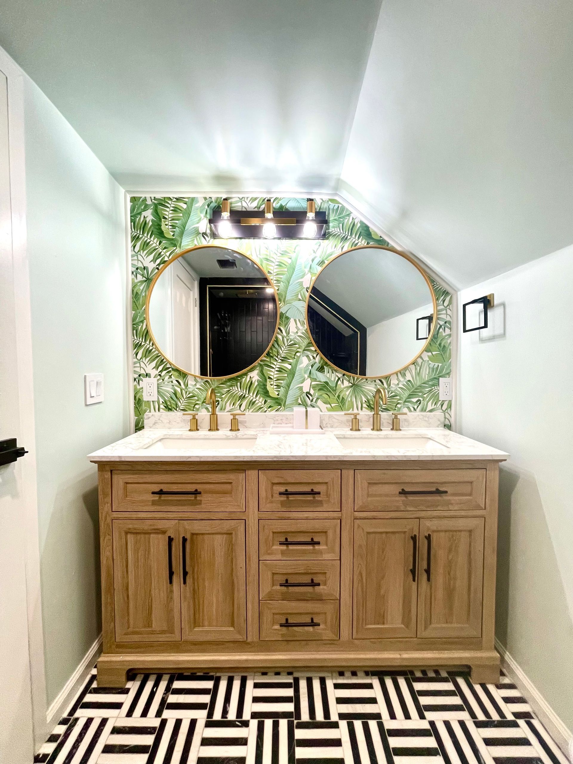 Bathroom with wooden vanity, round mirrors, and black and white floor tiles. Green and white patterned wallpaper.
