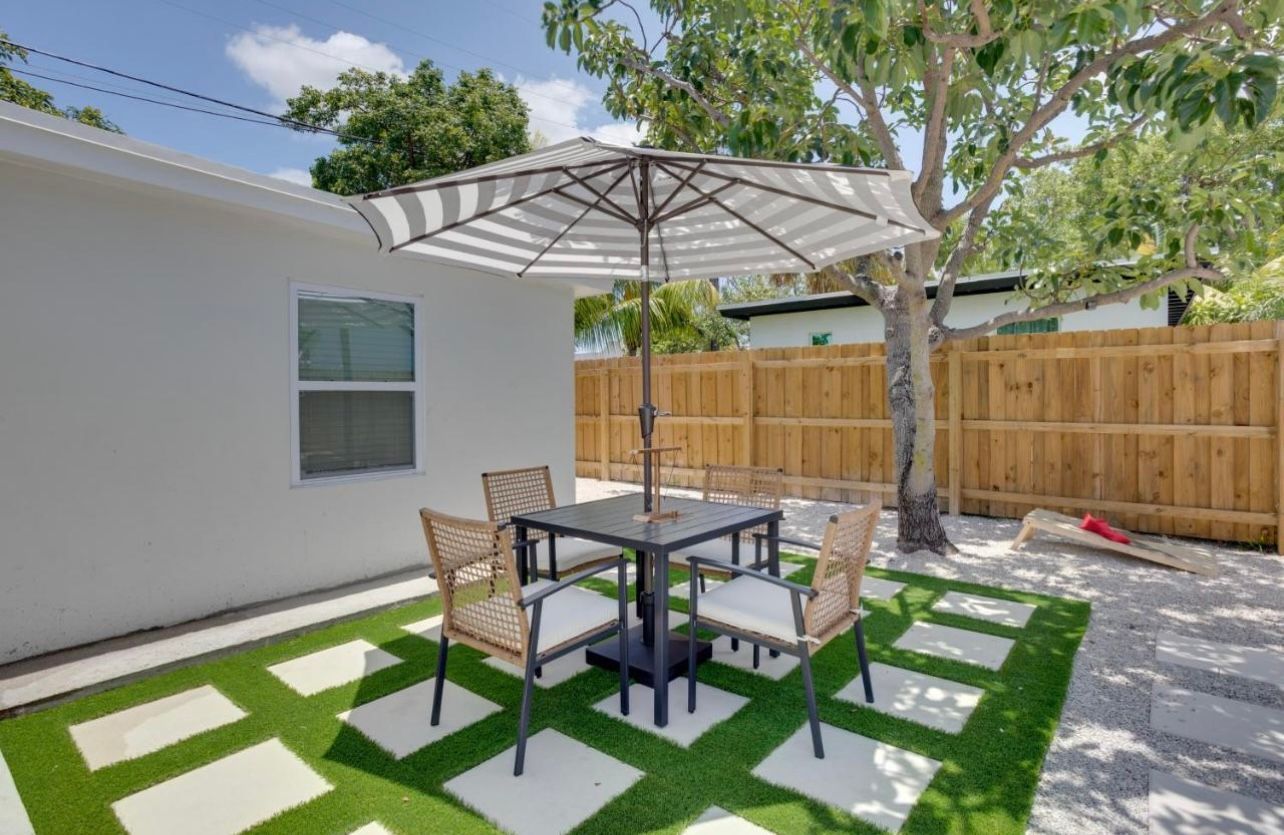 Backyard patio with table, chairs, and umbrella, on grass squares surrounded by rocks, near a fence.