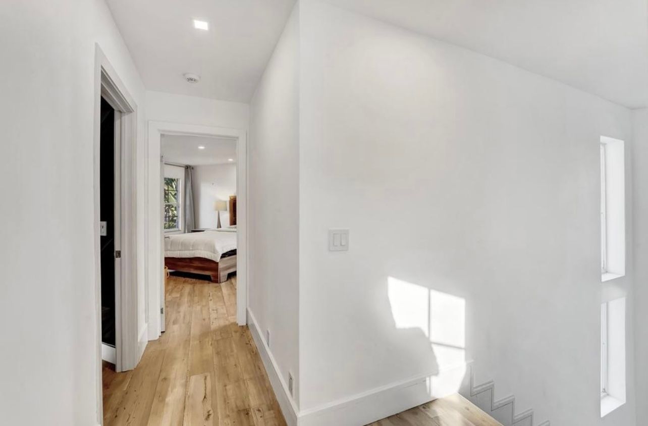 Hallway with white walls, hardwood floor, leading to a bedroom with sunlight.