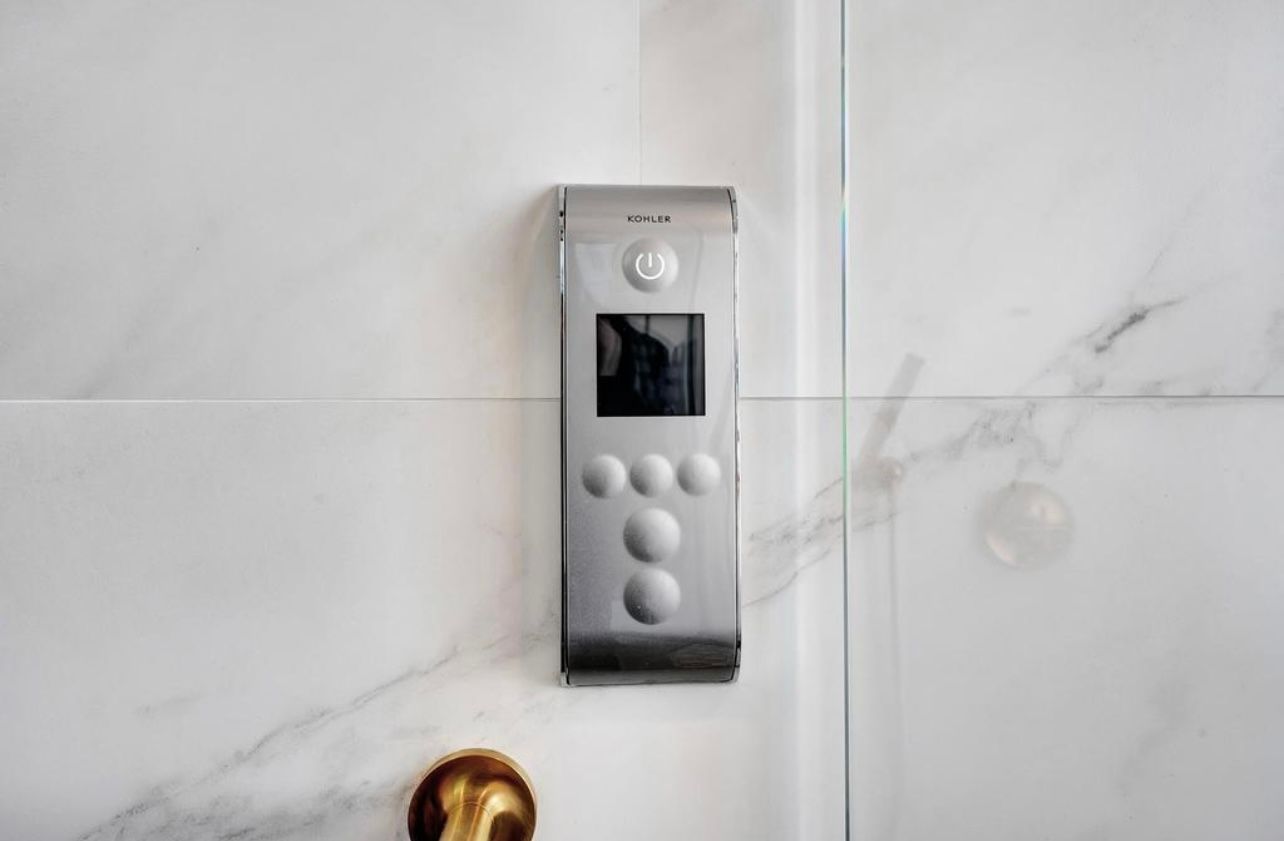 Silver control panel on a white tiled wall with a small screen and several round buttons.